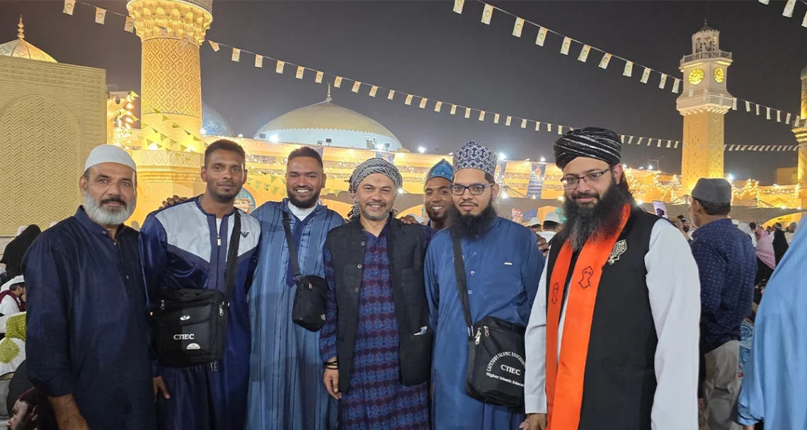 A group of South Africans stands with Hazrat Moulana Sayed Imraan Ziyaee(right) at the Urs of Shaykh Abdul Qadir Jilani (RA). They are pictured at night, with the illuminated mazaar and minarets in the background. (Photo: Ahlus Sunnah Media Network)